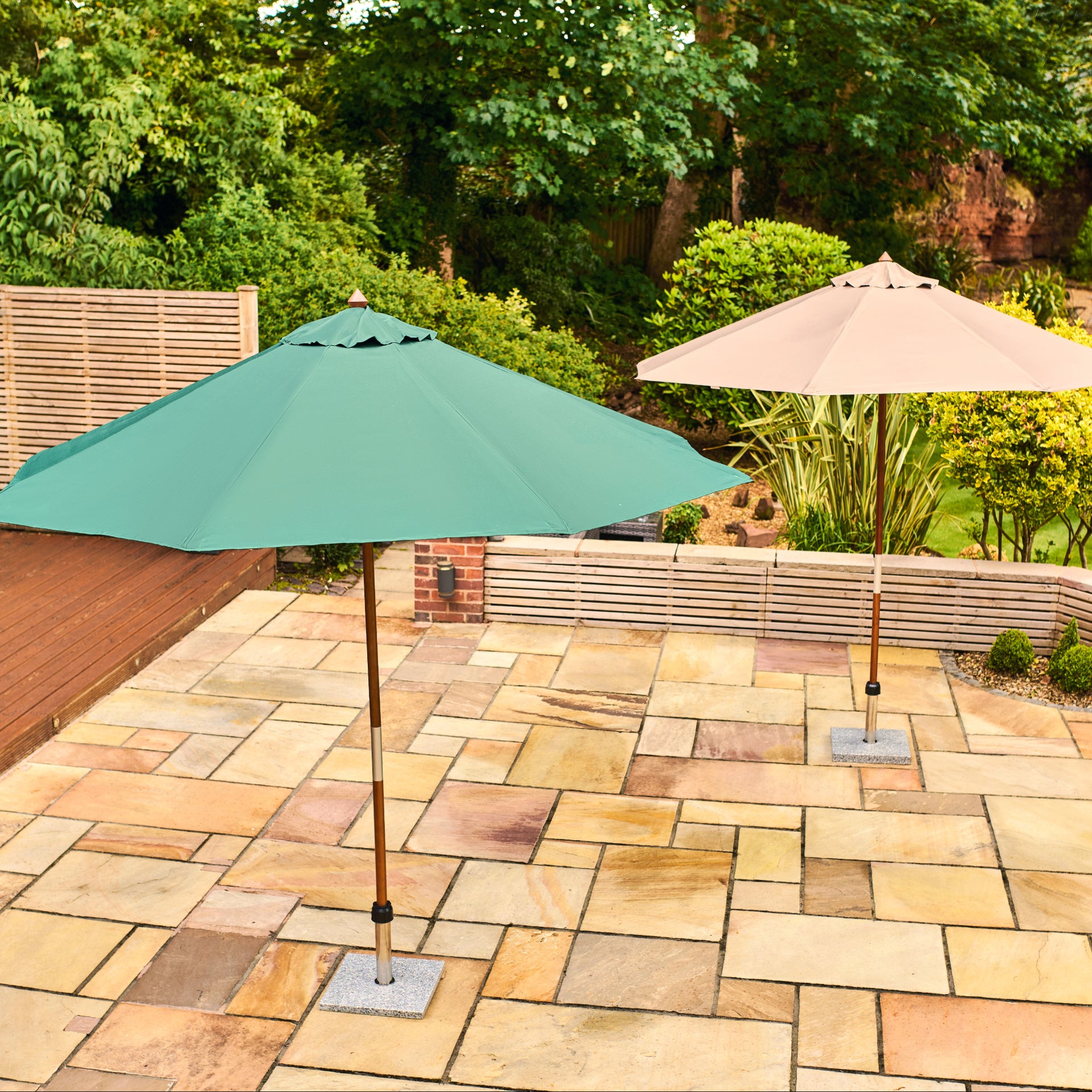 Two patio umbrellas on a stone patio in a landscaped garden. The foreground umbrella is teal green and the background umbrella is beige, both with square metal bases on a light-colored stone patio.