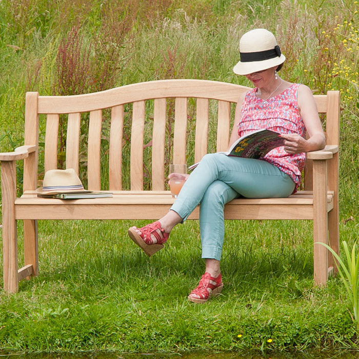 Woman sitting on a roble turnberry 5ft wooden bench in a garden reading a book.