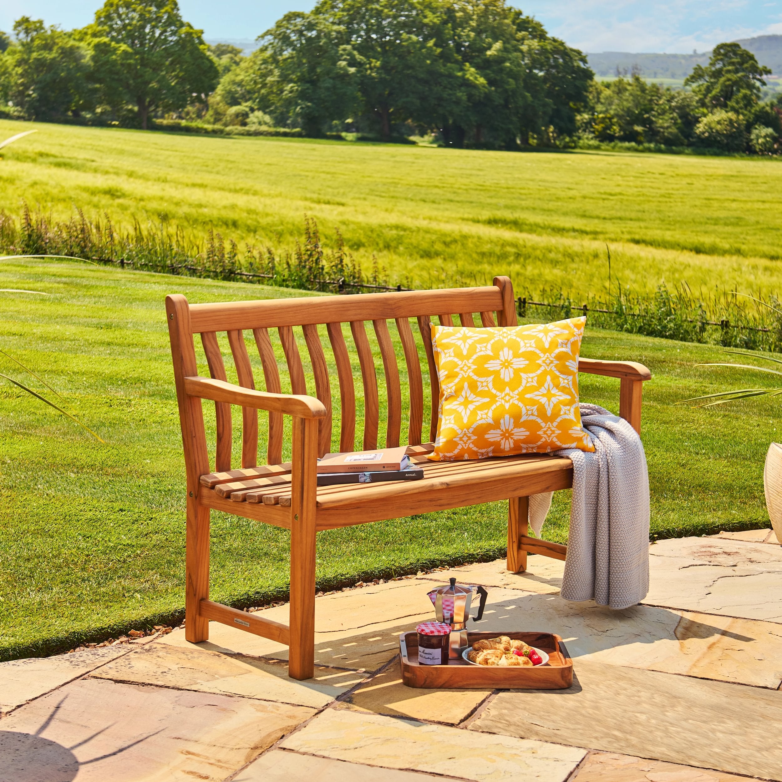 Wooden bench with a yellow cushion on a patio with a scenic view of green fields and trees.