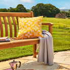 Wooden bench with a yellow patterned pillow and gray blanket in a garden setting