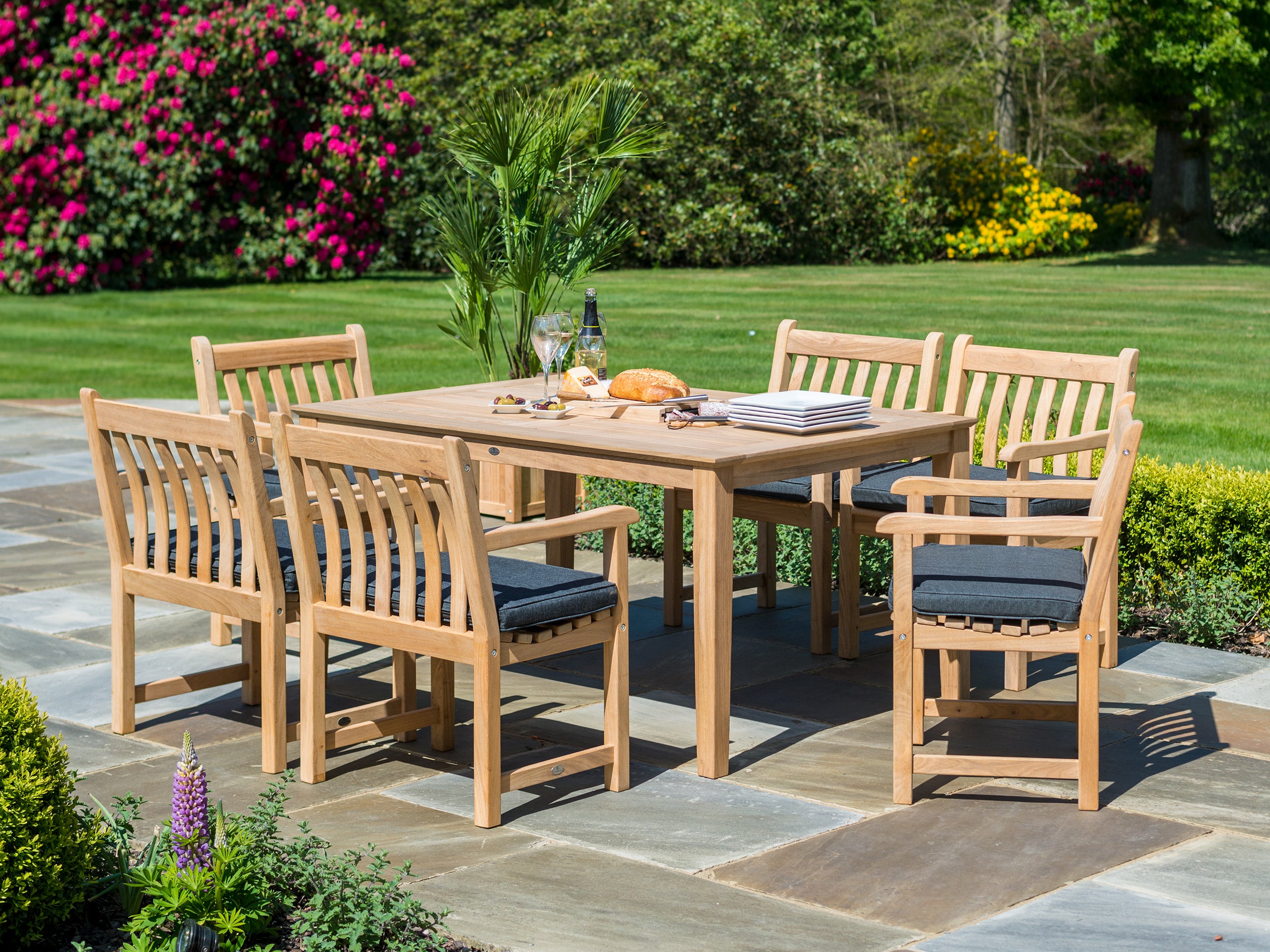 A 6-piece outdoor dining set consisting of a rectangular wooden table and six matching slatted armchairs with dark gray cushions, set on a stone patio in a lush green garden.