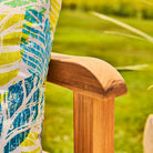 Close-up of a wooden chair arm with a colourful cushion on it, set against a blurred green outdoor background.