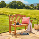 Wooden bench with a pink cushion and a hat on a stone patio with a scenic background of green fields and trees.
