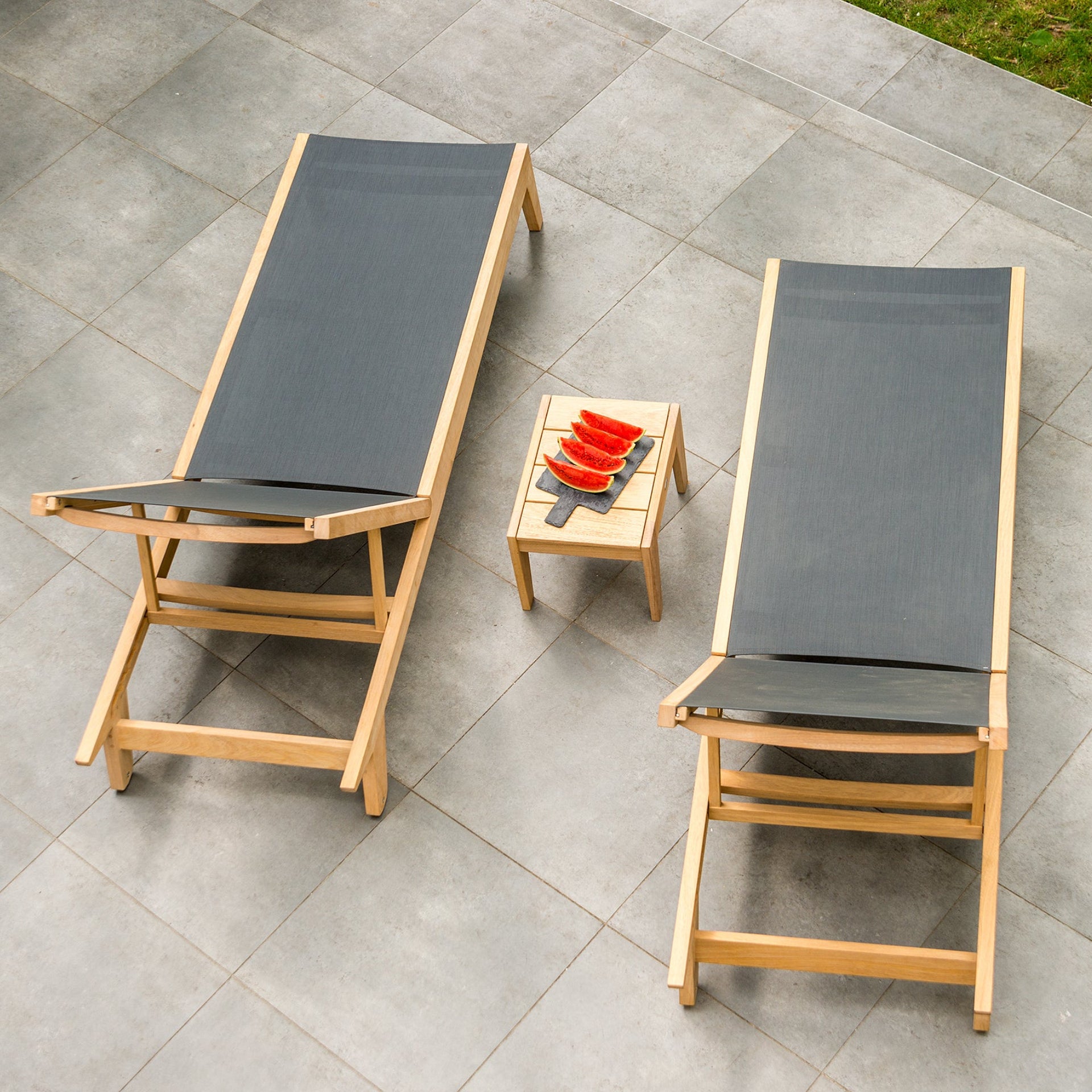 Two wooden lounge chairs with dark cushions on a tiled patio, accompanied by a small table with watermelon.