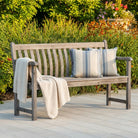 Wooden bench with striped pillows and a blanket on a wooden deck with greenery in the background