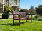 A mahogany wooden garden bench with slatted back and seat, placed on a green lawn in front of a stone building with shrubs and blue sky.