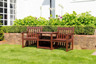 A three-piece mahogany outdoor wooden garden set consisting of two armchairs and a small central table, arranged on a green lawn in front of a brick planter and a white house.