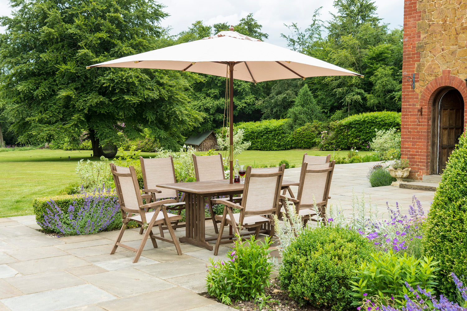 Garden setting with wooden dining table and sling recliner chairs under a large parasol, surrounded by greenery.