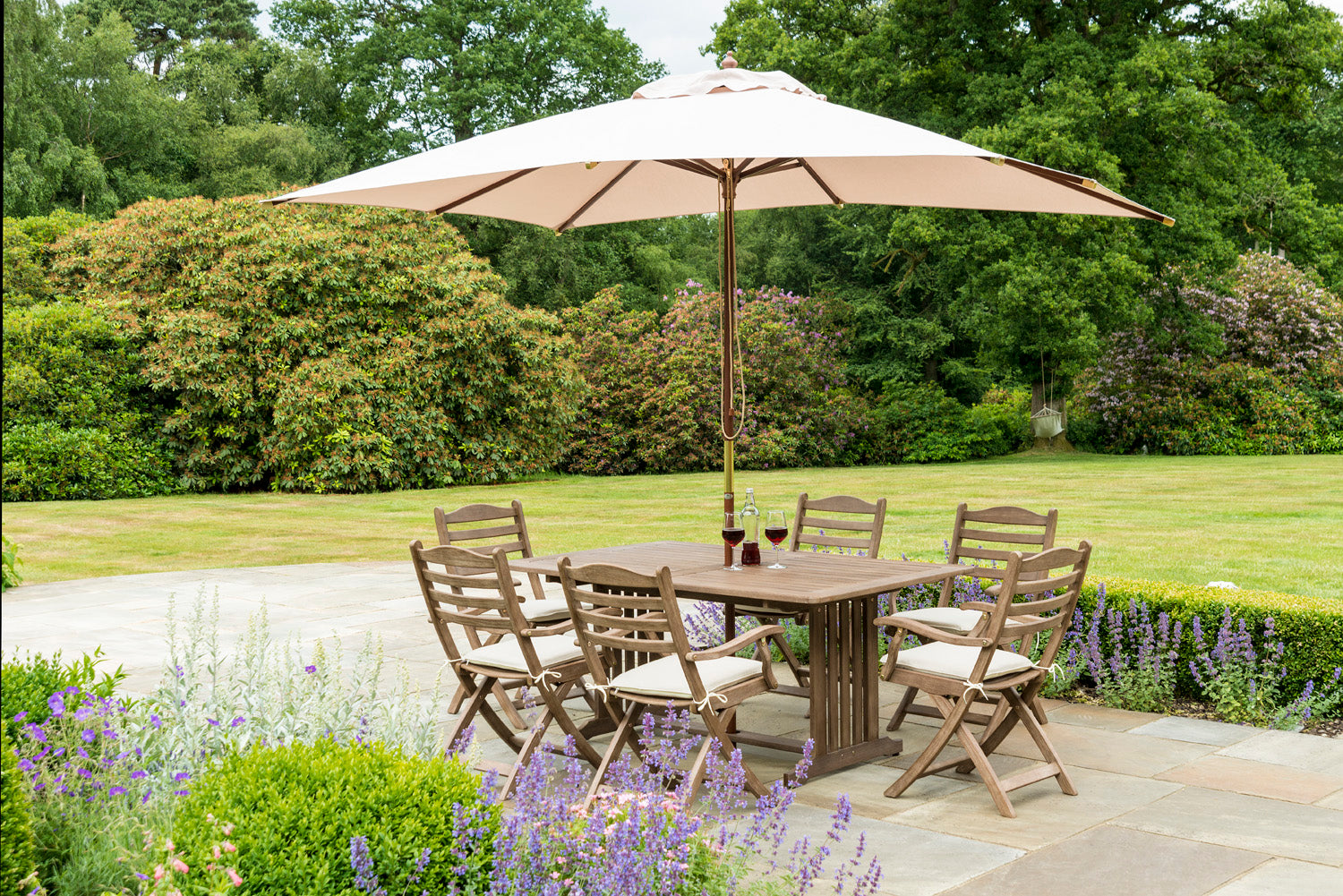 Garden setting with wooden dining table and chairs under a large beige umbrella, surrounded by greenery and flowers.