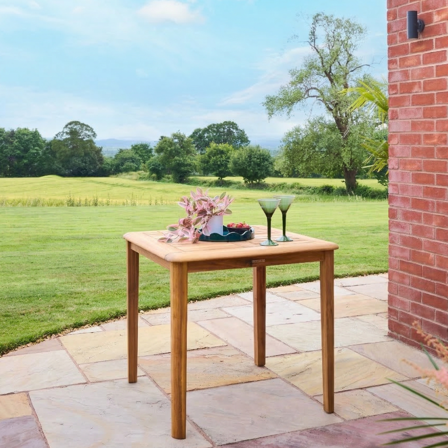 Wooden table with flowers and glasses on a stone patio with a grassy field and trees in the background.