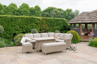 Outdoor patio furniture set with a curved sectional sofa, matching armchair, ottoman, and a rectangular wicker coffee table, arranged on a stone patio in a landscaped garden with a hedge and a small gazebo.
