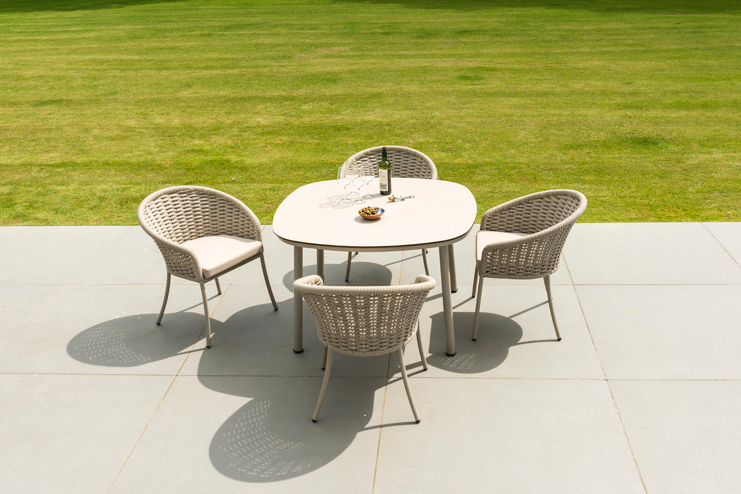 Outdoor patio set with a round table and four rope-woven chairs on a tiled patio with grass in the background.