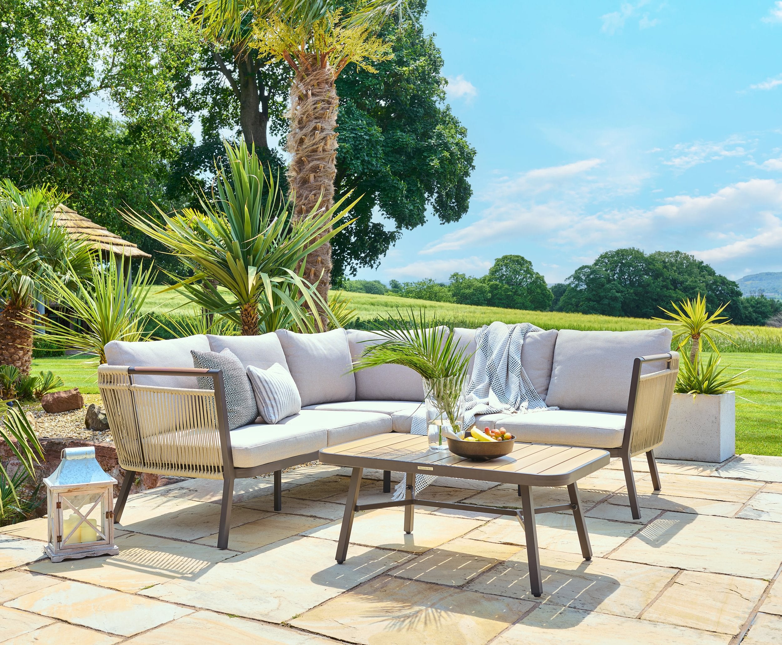 Outdoor patio set with sectional sofa, chairs, and coffee table on a stone patio with greenery in the background.