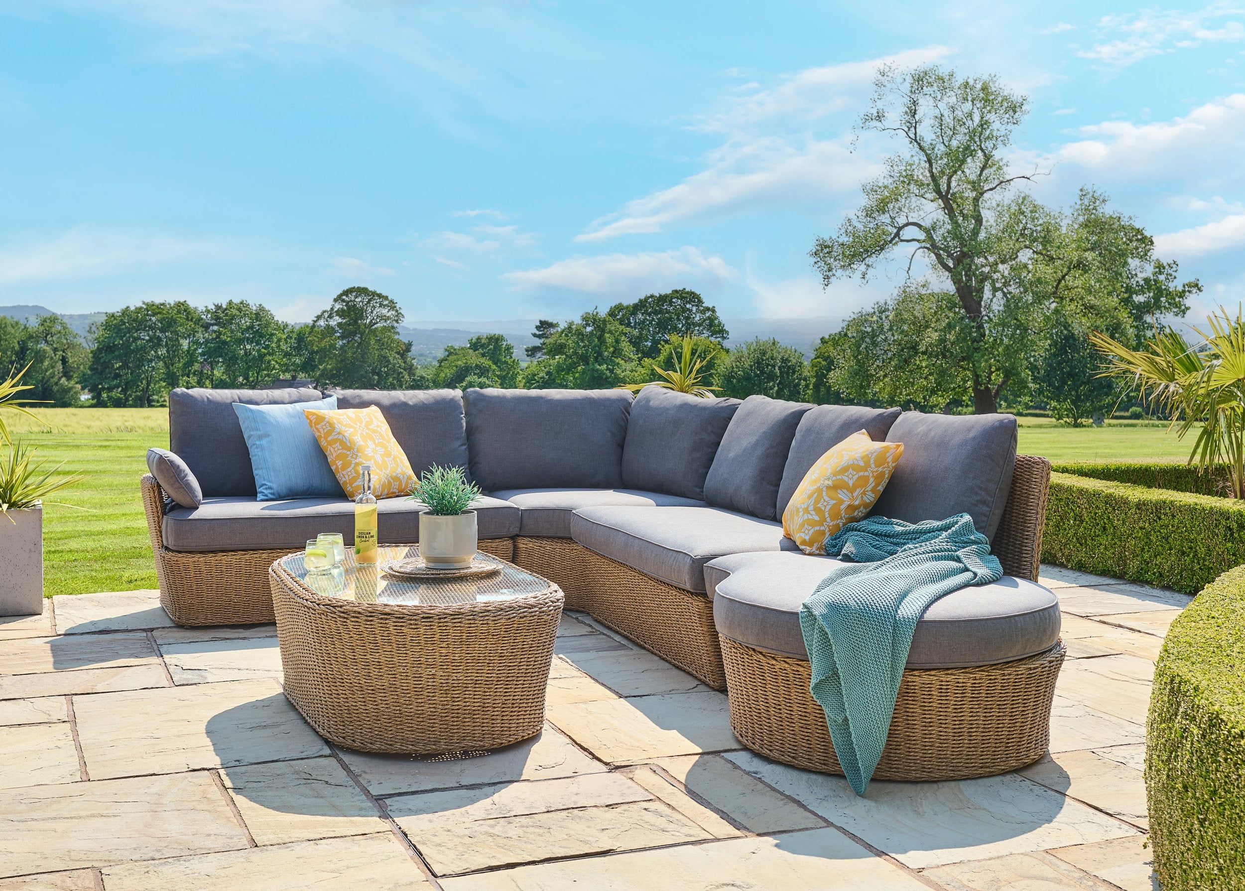 Outdoor patio furniture set with a sectional sofa and round table on a stone patio with a scenic background.