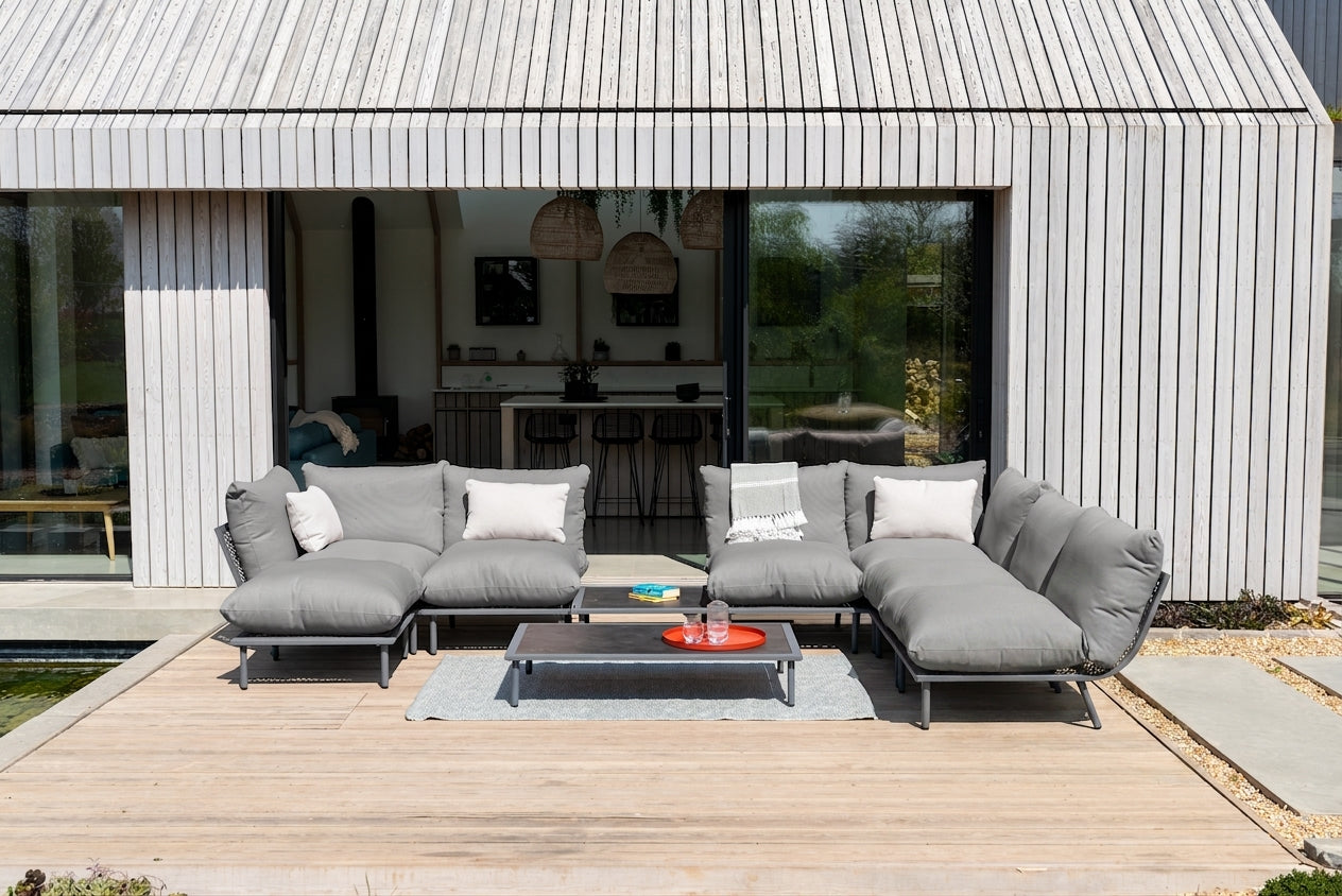 A modern outdoor sectional sofa set in gray fabric with white accent pillows, arranged on a wooden deck in front of a contemporary house with vertical siding and large glass windows.