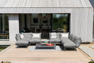 A modern outdoor sectional sofa set in gray fabric with white accent pillows, arranged on a wooden deck in front of a contemporary house with vertical siding and large glass windows.