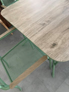 A close-up of a dining table with a light gray wood-grain top and mint green metal legs, alongside matching mint green mesh chairs.