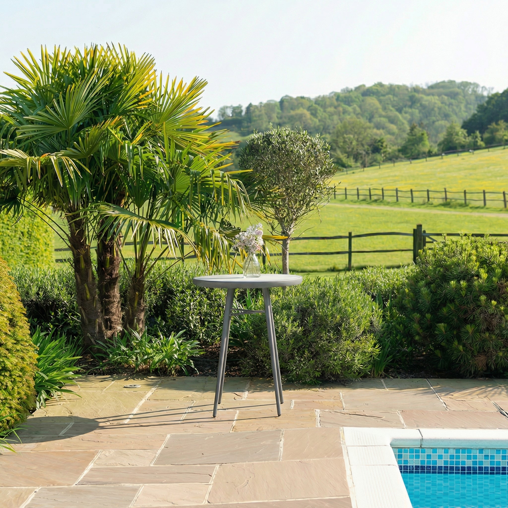 Outdoor setting with a table by a pool, surrounded by greenery and a fence.