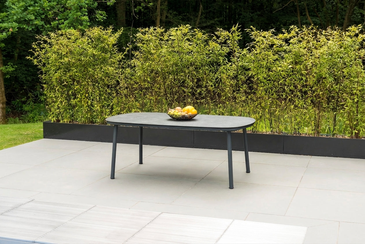 Outdoor patio table with pebble grey top. Rectangular with curved corners, on a tiled patio area with greenery in the background and a bowl of fruit in the centre