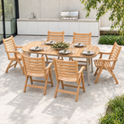 An outdoor dining set consisting of a rectangular teak wood table and six matching slatted armchairs, arranged on a concrete patio in a backyard setting with a grill and greenery in the background.
