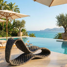A modern wavy lounge chair made of rope dark gray rope sits beside an infinity pool overlooking a turquoise sea and distant mountains. Palm trees, potted olive trees, and cream patio umbrellas frame the scene.