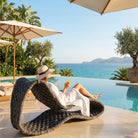 A woman relaxes on a curved rope lounge chair beside an infinity pool, with the sea and tropical trees in the background. She is wearing a white dress and a wide-brim straw hat and appears to be holding a drink.