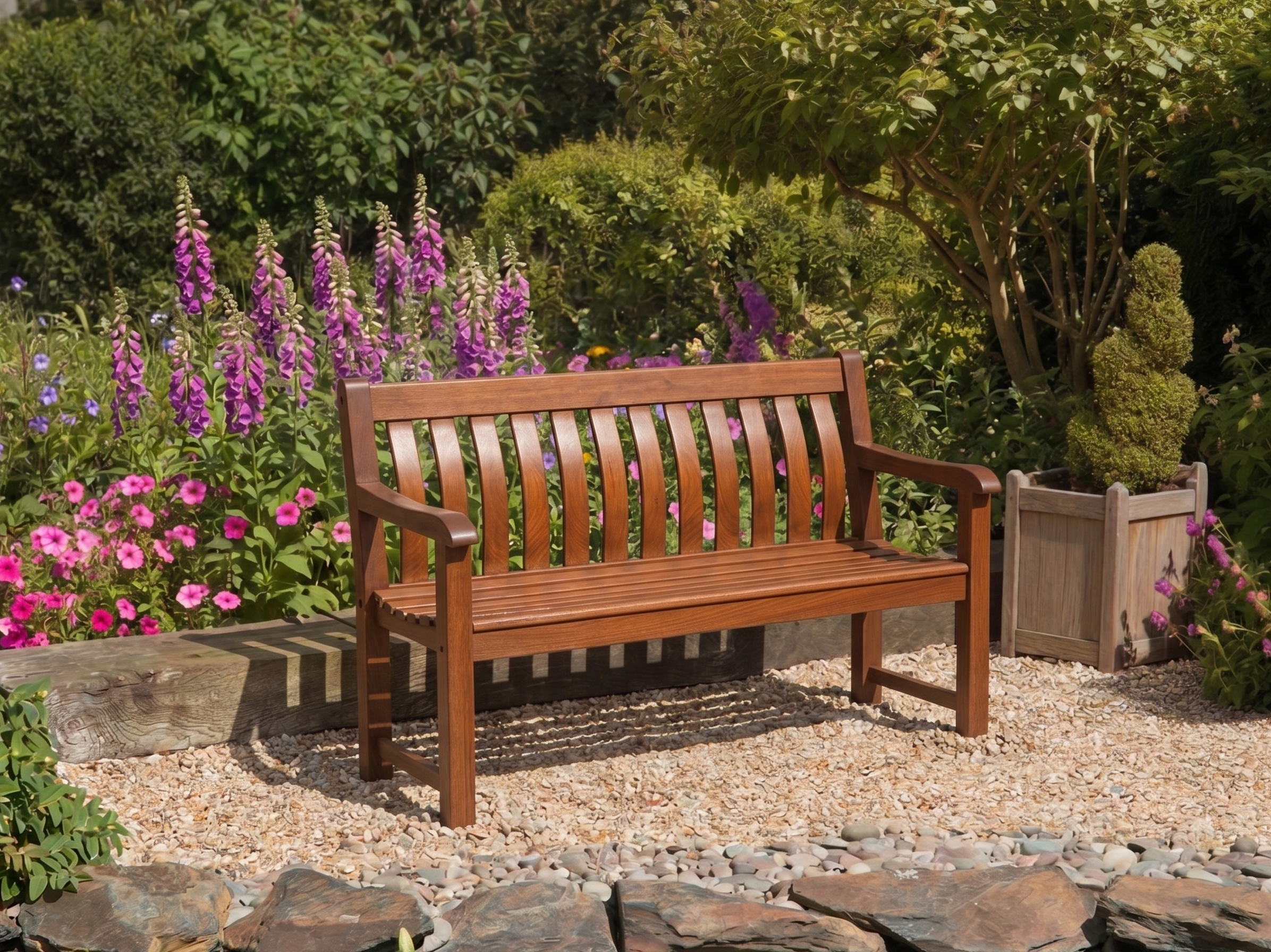 A wooden garden bench in mahogany with a slatted back and seat, placed on a gravel path in a landscaped garden with purple flowers and green foliage.