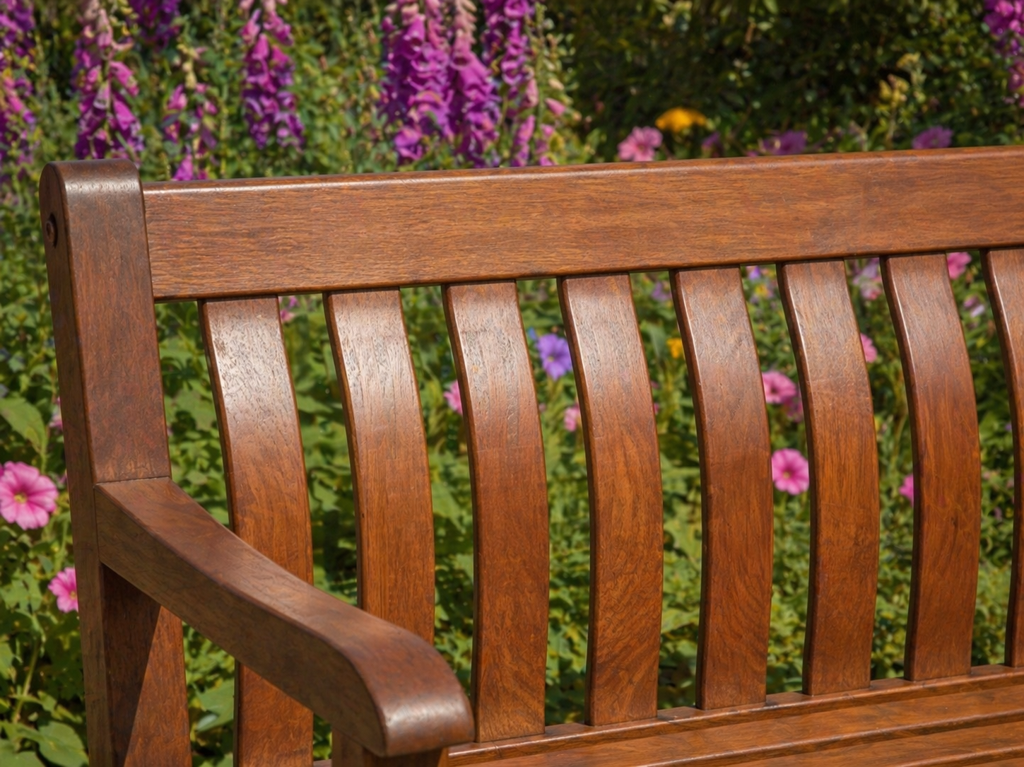 A close-up of a wooden garden bench in mahogany, set against a blurred background of green foliage and purple flowers.