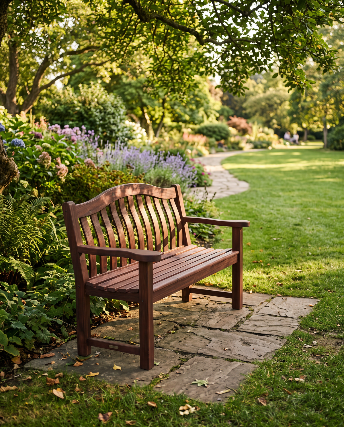 A dark brown wooden garden bench with slatted back and seat, placed on a stone path in a lush green park with flowers and trees in the background.