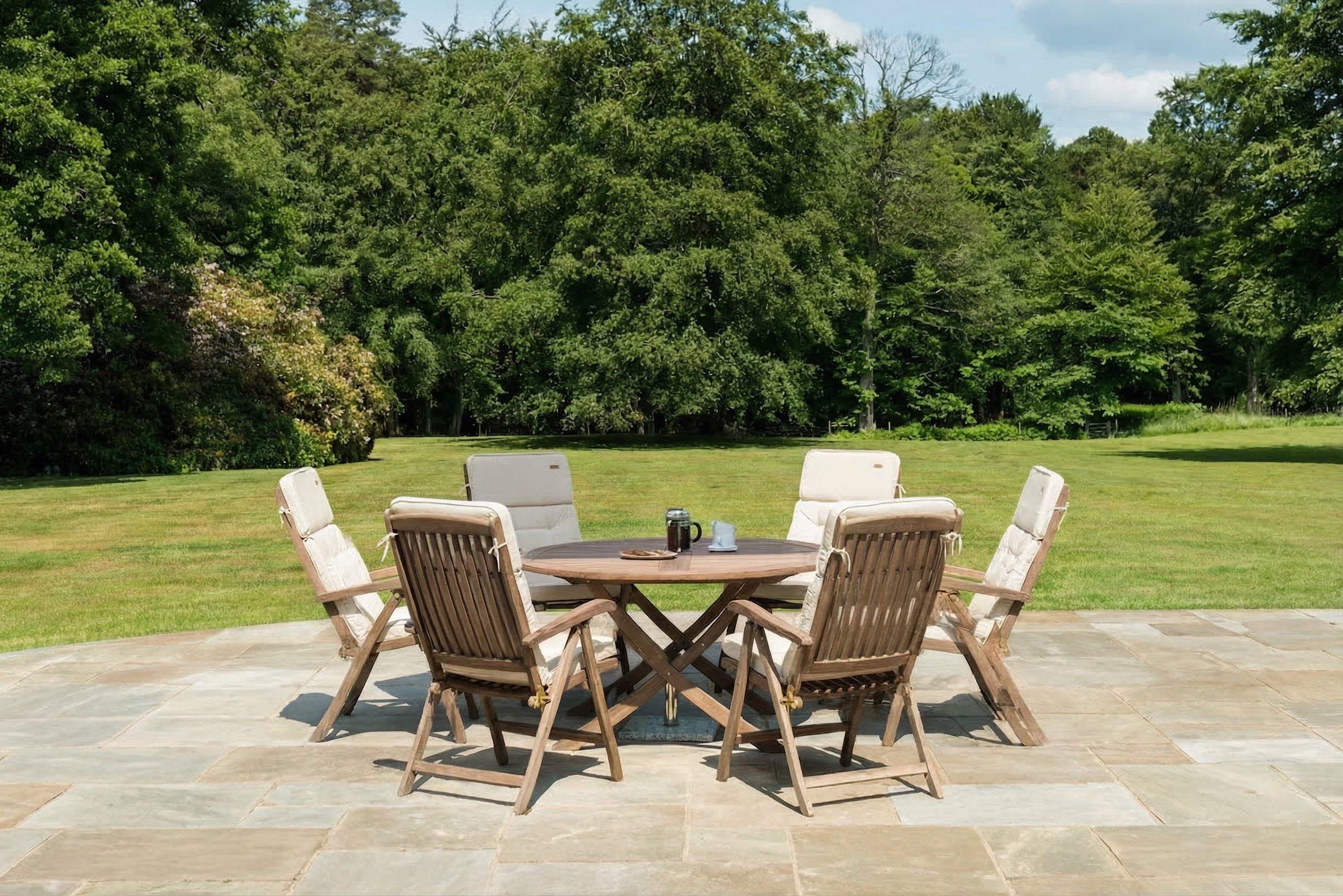 Outdoor patio set with wooden table and chairs on a stone patio with green grass and trees in the background.