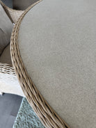 Close-up of a round outdoor dining table with a light beige woven wicker frame and a thick, textured beige fabric cushion.