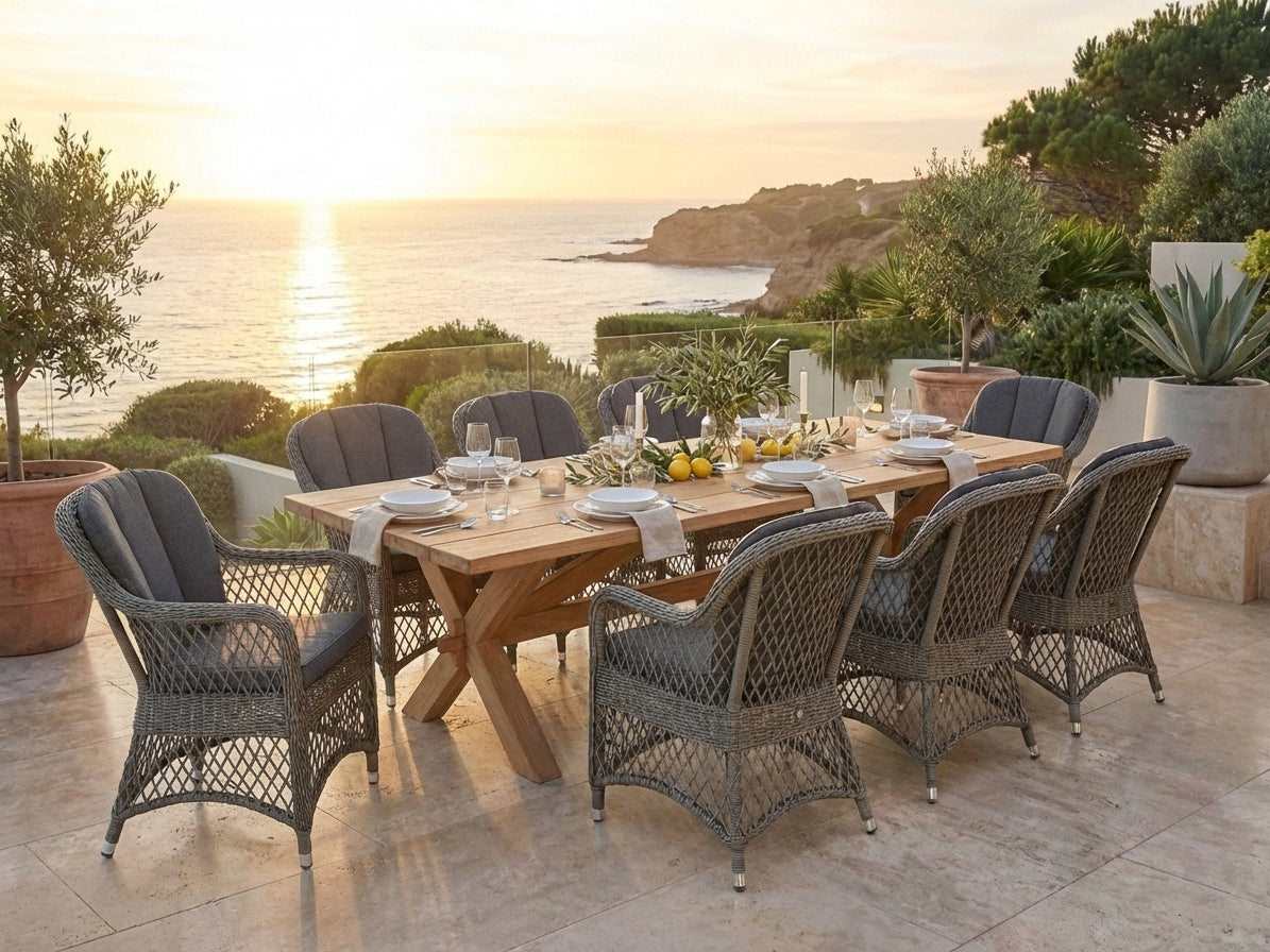 Outdoor dining setup with a wooden table and wicker chairs by the ocean during sunset.