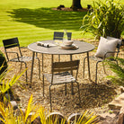 Garden setting with a round metal table and chairs on a gravel area, surrounded by grass and plants.