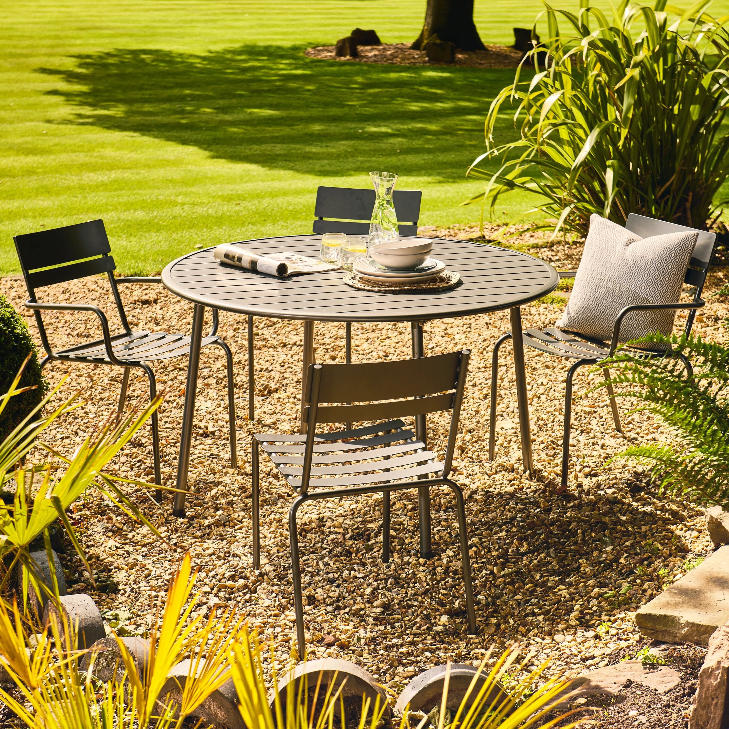 Garden setting with a round metal table and chairs on a gravel area, surrounded by grass and plants.