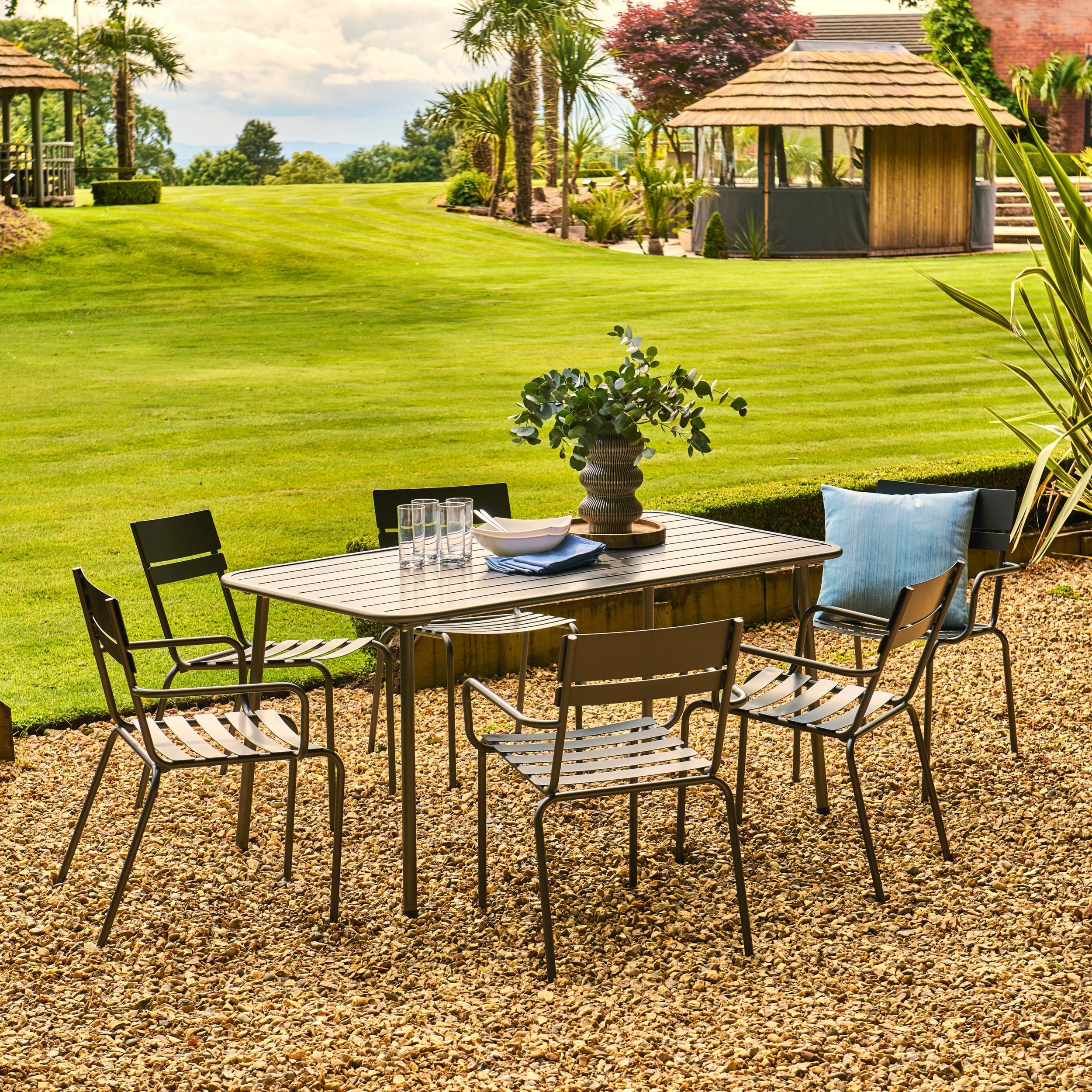 Outdoor patio set with table and chairs on a gravel area, grassy field, and gazebo in the background.