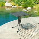 Black mesh table on a wooden deck by a pool with greenery in the background