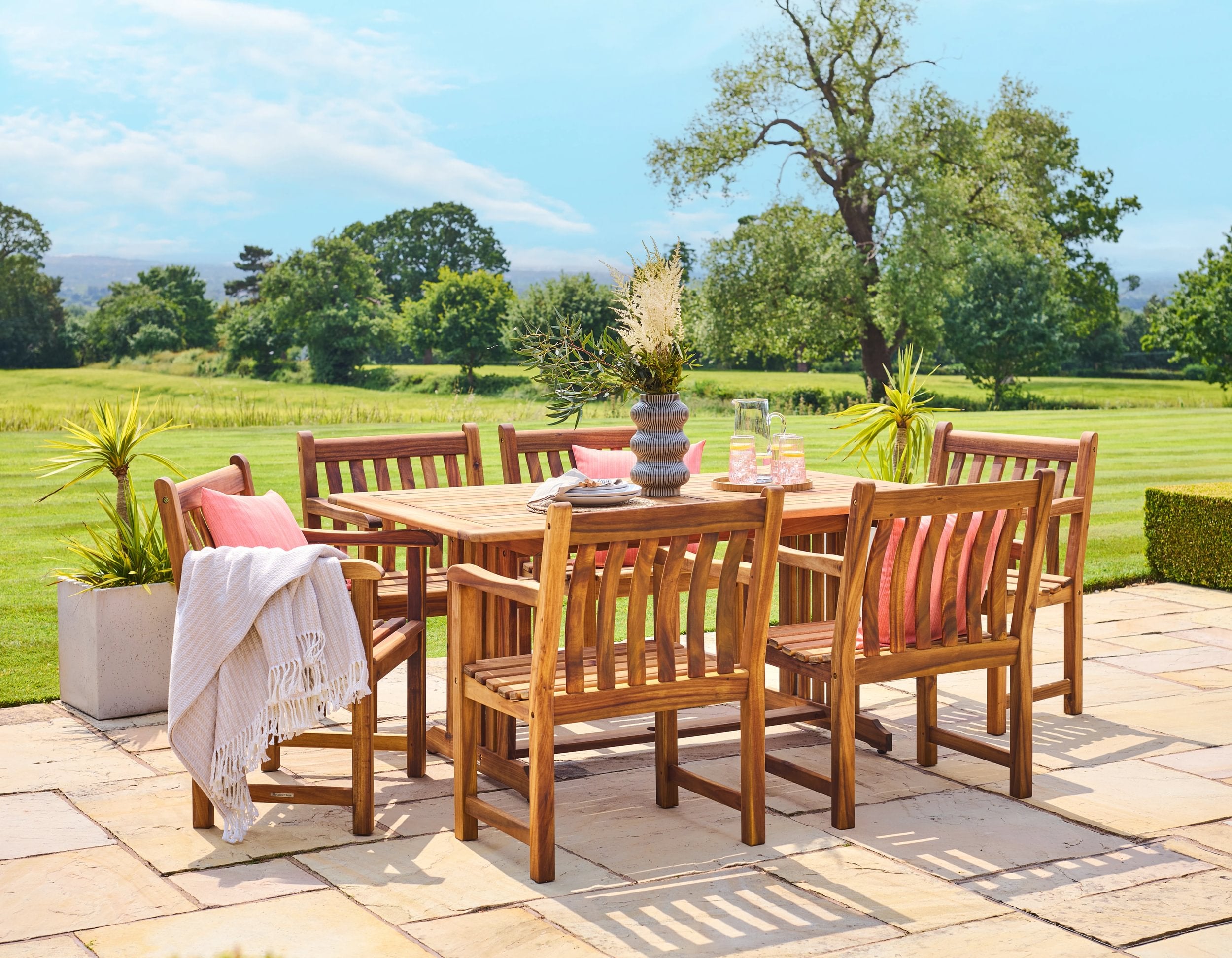 Outdoor dining set with wooden table and chairs on a patio, surrounded by greenery.
