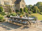 Outdoor dining setup with a large table and chairs on a stone patio in a garden setting.