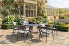 A 7-piece outdoor dining set consisting of a round dark grey table and six matching chairs with gray slatted backs and light blue cushions, arranged on a stone patio in front of a stone house with a glass conservatory.