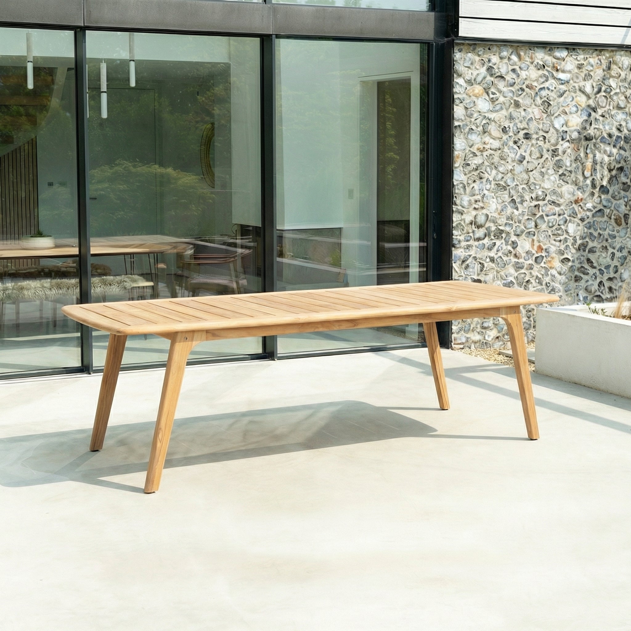 Wooden outdoor dining table on a patio with a modern building and stone wall in the background.