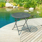 Black metal folding table on a wooden deck by a pond with greenery.