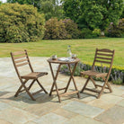 Wooden outdoor table with two chairs on a stone patio with greenery in the background