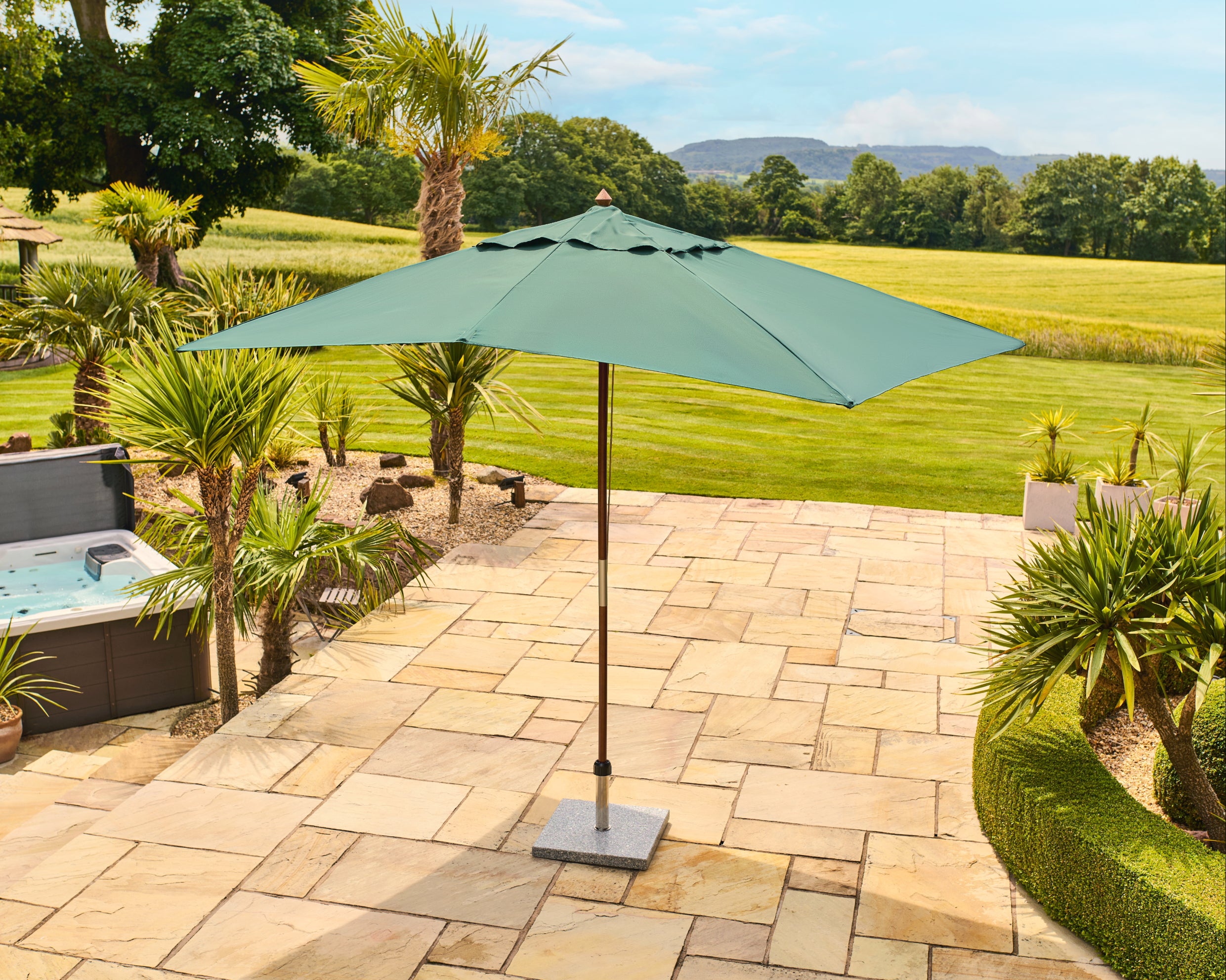 A green patio umbrella with a square canopy and central pole, standing on a stone patio in a landscaped backyard with grass, trees, and a hot tub nearby.