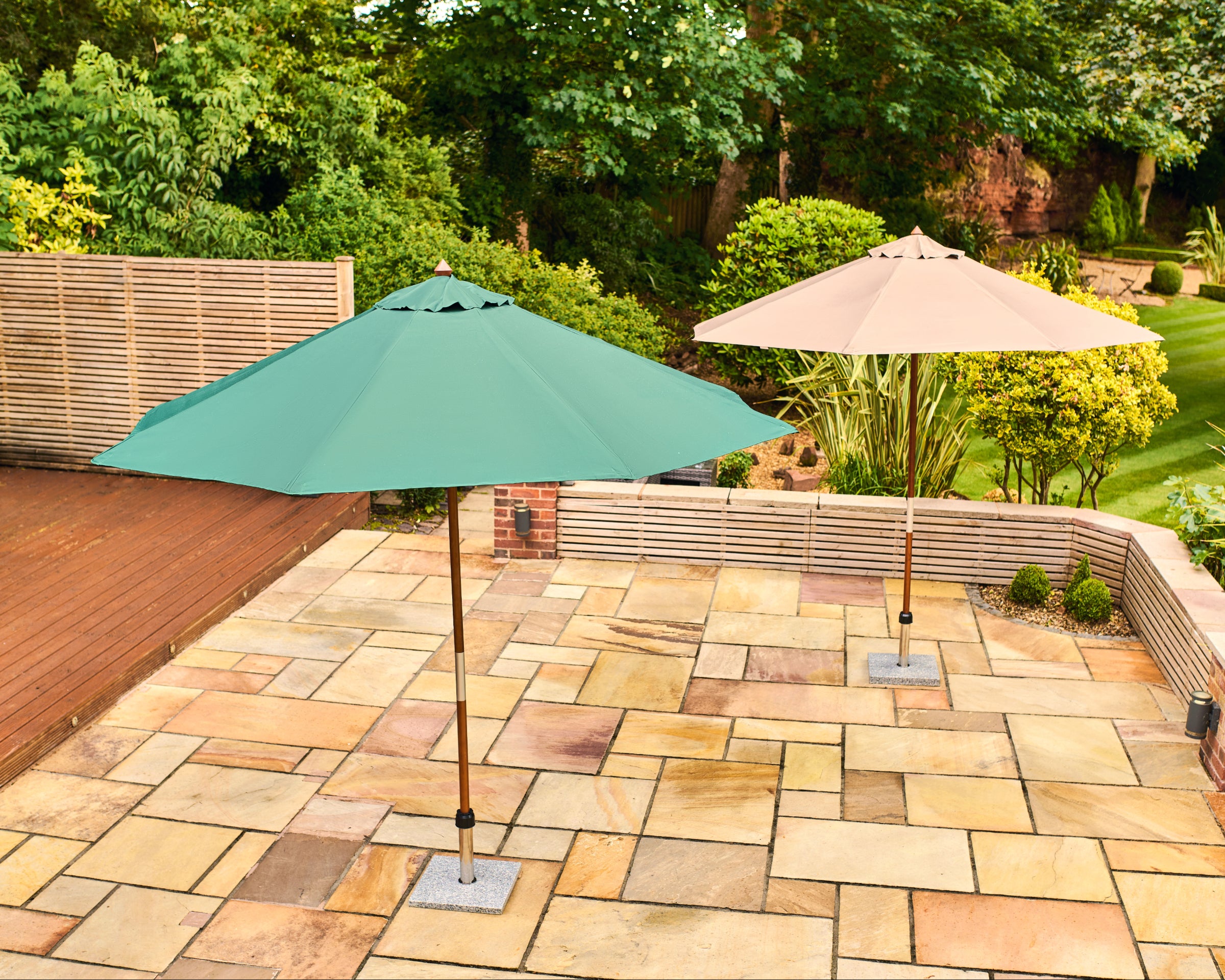 Two patio umbrellas on a stone patio in a landscaped garden. The foreground umbrella is teal green and the background umbrella is beige, both with square metal bases on a light-colored stone patio.