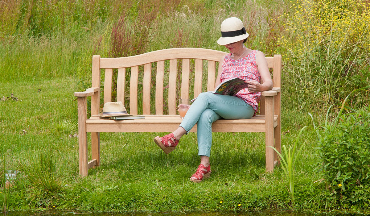 Woman sitting on a roble turnberry 5ft wooden bench in a garden reading a book.